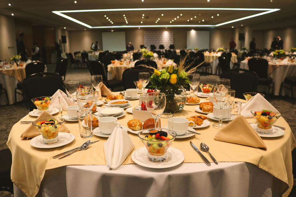Elegant banquet hall setup with round tables covered in tablecloths, neatly folded napkins, glassware, and plated fruit cups, prepared for an event.