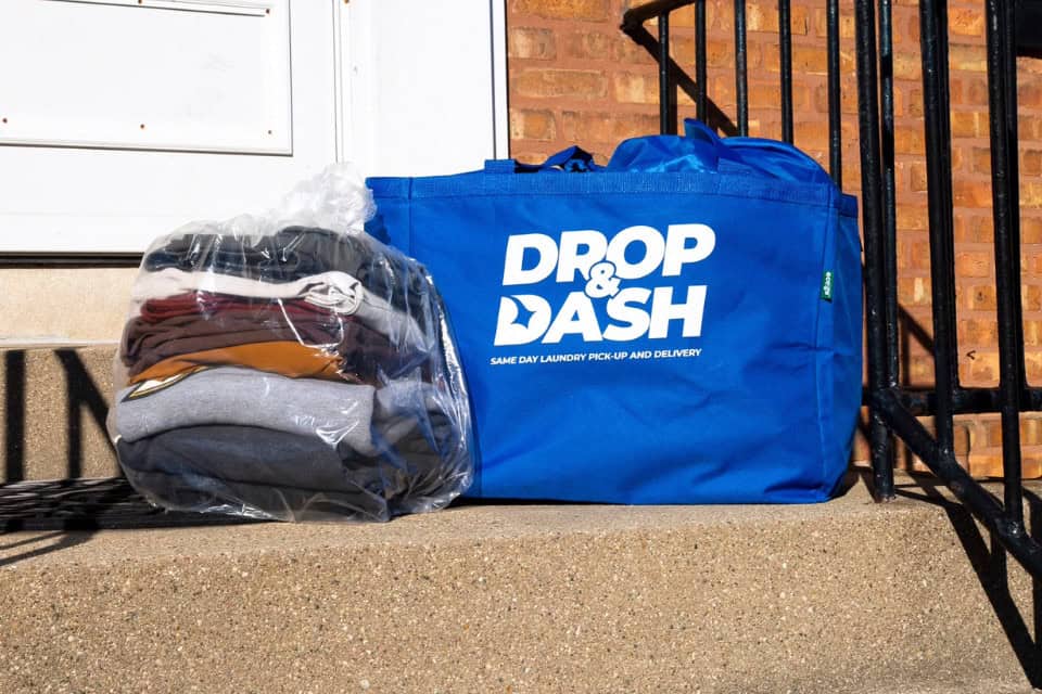 Folded laundry in a plastic bag next to a blue Drop & Dash tote bag on a doorstep, freshly delivered to a customer.