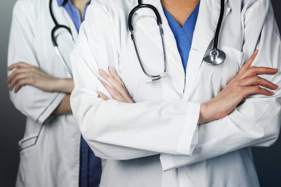 Close-up of two healthcare professionals in clean white lab coats with stethoscopes, arms crossed.
