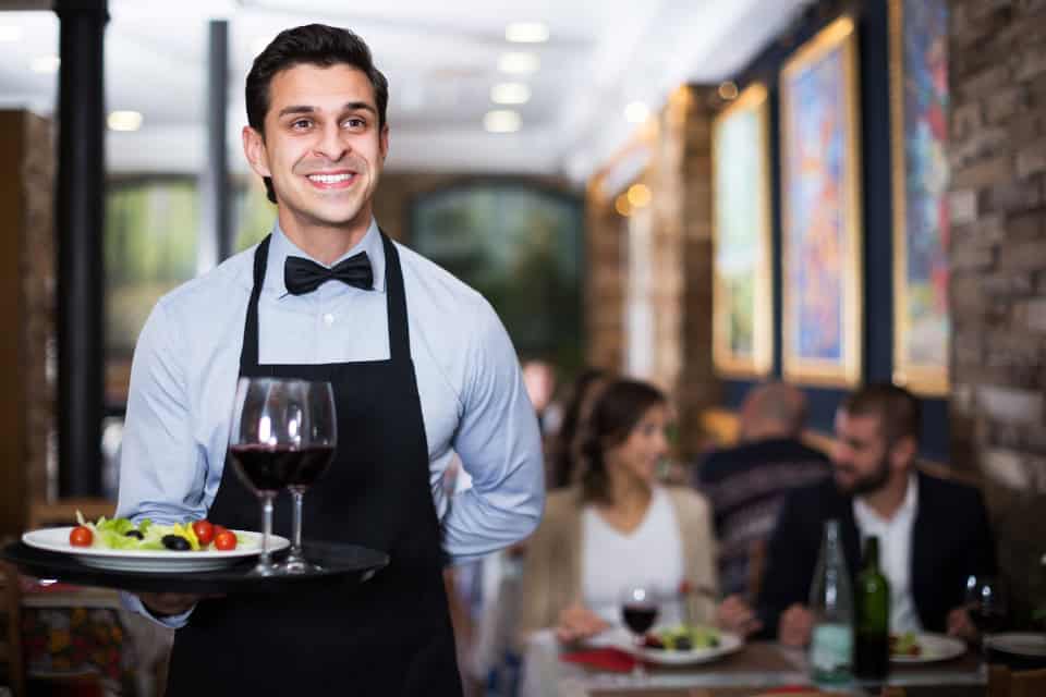 Server wearing a clean black apron and bow tie delivering food and wine at a Chicago restaurant, representing Drop & Dash commercial laundry service for restaurants.