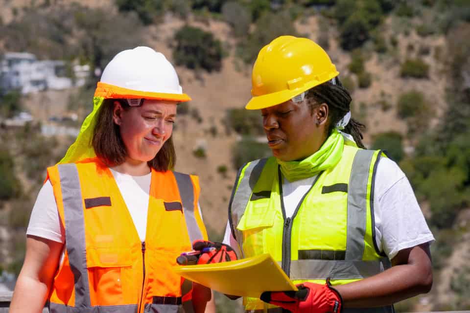 Two construction workers wearing reusable high-visibility safety vests and hard hats reviewing documents outdoors