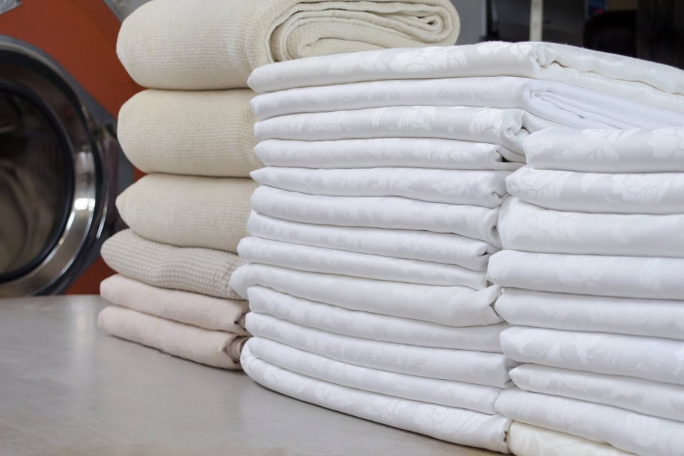 Freshly folded commercial linens stacked on a laundry table near industrial washers.